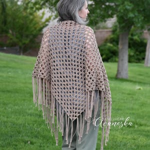 May include: A light brown triangular crochet shawl featuring an openwork diamond pattern and long fringe along the bottom edges. The garment is draped over the shoulders and back, showcasing the intricate yarn texture and the knotted fringe detail.