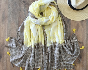 May include: A rectangular scarf featuring a bright yellow upper section and a taupe lower section with geometric patterns, finished with small yellow tassels along the edges, accompanied by a straw hat with a black band and the text Avaneska MODERN KNITWEAR.