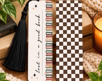 May include: A rectangular bookmark with a black tassel, featuring a cream background, a vertical row of colorful illustrated books, and the text "lost in a good book" with star accents. The reverse side has a brown and white checkered pattern.