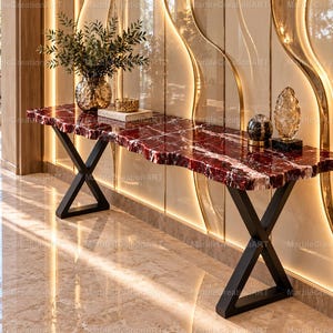 May include: A long console table featuring a deep red, polished marble slab top with natural, uneven live edges. The table is supported by two black metal X-shaped legs. Decorative items including a glass vase with greenery, a small box, and two sculptural objects are placed on the surface.