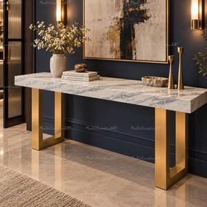 May include: A rectangular console table featuring a white marble top with grey veining and two brushed gold metal U-shaped legs. The table surface holds a white vase with white flowers, two stacked books, a small bowl, and two gold candlesticks.