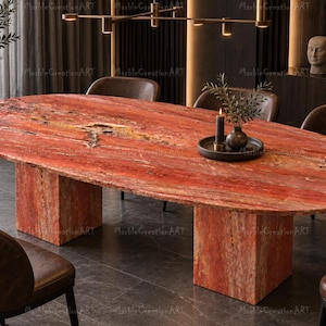 Pode incluir: Uma mesa de jantar oval com um tampo de pedra natural vermelho vibrante com veios e textura visíveis. A mesa é suportada por dois pedestais retangulares espessos feitos da mesma pedra vermelha, criando uma peça de mobiliário sólida.