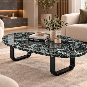 Op de afbeelding: Een ovale salontafel met een gepolijst donkergroen marmeren blad met witte aders en twee zwarte U-vormige metalen poten. Op het tafeloppervlak staan een kleine beige kom, een beige vaas met groen blad en twee op elkaar gestapelde boeken.