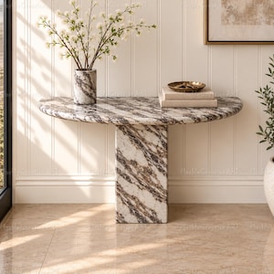May include: A semi-circular console table crafted from white and brown veined marble, featuring a thick central pedestal base. A matching cylindrical marble vase holds white flowers, and a brass bowl sits atop two stacked rectangular books.