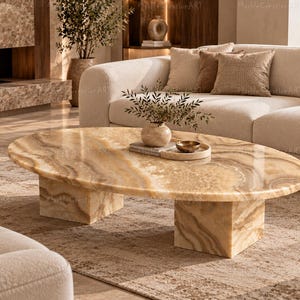May include: An oval coffee table crafted from polished beige and brown veined marble, featuring a smooth tabletop surface supported by two thick, rectangular block-style legs made of the same matching stone material.