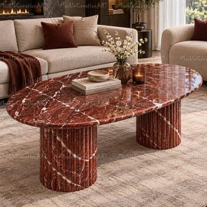 May include: An oval coffee table made of deep red marble with white veining. The table features a smooth, polished top surface and two cylindrical, fluted bases. The surface holds a small bowl, two stacked books, a glass vase with flowers, and a candle.