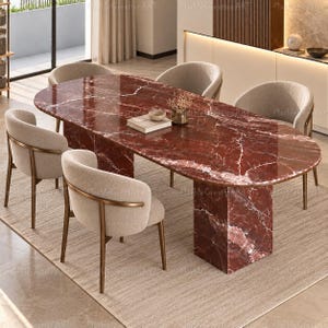 Pode incluir: Uma mesa de jantar oval com um tampo de mármore vermelho profundo polido e uma base de pedestal de mármore a condizer, rodeada por quatro cadeiras estofadas em bege com encostos curvos e estruturas metálicas finas, com um livro e um vaso.
