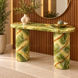 May include: An oval console table crafted from green onyx stone with a polished finish. The table features a smooth top and two cylindrical, fluted bases with diagonal veining patterns in shades of green, white, and amber.