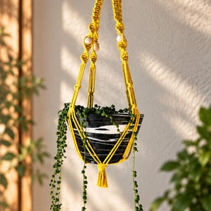 May include: A bright yellow macrame plant hanger featuring knotted cord and light wooden beads, suspending a black and white marbled bowl containing a trailing string of pearls plant with long, thin green stems and small round leaves.
