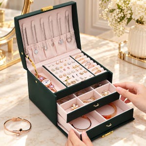 Puede incluir: Un joyero de color verde oscuro con interior rosa claro, que cuenta con una tapa superior con ganchos para collares, un compartimento para reloj, ranuras para pendientes y anillos, y dos cajones extraíbles con divisores internos.
