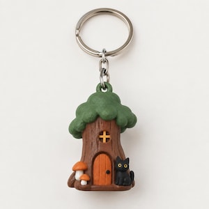 May include: A keychain featuring a brown tree trunk house with a green leafy top, a small orange arched door, a yellow window, two orange mushrooms at the base, and a small black cat sitting next to the door, all attached to a silver metal ring.