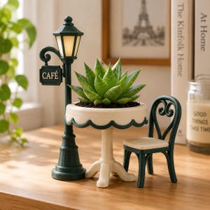 May include: A miniature dark green street lamp with a light-up top and a sign reading CAFÉ, paired with a cream-colored round table holding a green succulent plant and a matching dark green chair with a heart-shaped backrest design.