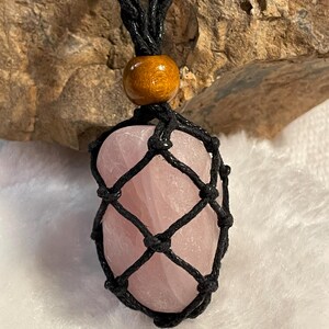 May include: A polished, light pink rose quartz stone encased in a black macrame cord netting. The pendant hangs from a braided black cord featuring a single, smooth, round, honey-colored wooden bead positioned directly above the stone netting.