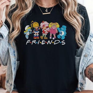 Könnte beinhalten: Ein schwarzes Kurzarm-T-Shirt mit einem Grafikdruck von fünf bunten Zeichentrickfiguren, die in einer Reihe über dem weißen Schriftzug FRIENDS mit bunten Punkten zwischen den Buchstaben stehen. Die Figuren umfassen einen Schlumpf, Rainbow Brite, Erdbeertörtchen, eine Cabbage Patch Puppe und einen blauen Glücksbärchi.