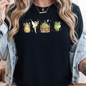 May include: A black short-sleeved t-shirt featuring a horizontal graphic print of a brown rabbit wearing a leaf hat, a small fairy with wings, a whimsical cottage covered in foliage, and a large green moth, all surrounded by small yellow stars.