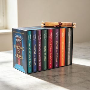 以下が含まれることがあります： サラ・J・マース著『Throne of Glass』のハードカバー本8冊セット。ティール、ブルー、パープル、グリーン、レッド、バイオレット、オレンジ、ブラックのカラフルな背表紙が特徴。茶色のリボンで結ばれた巻物が上に置かれている。