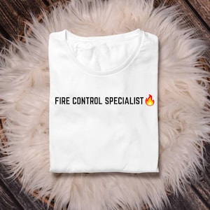 Könnte beinhalten: Ein weißes kurzärmeliges T-Shirt mit dem schwarzen Schriftzug "FIRE CONTROL SPECIALIST" auf der Brust, gefolgt von einem kleinen Flammen-Emoji. Das Kleidungsstück hat einen Rundhalsausschnitt und ist ordentlich gefaltet.