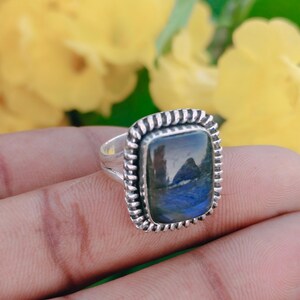 May include: A silver ring featuring a rectangular labradorite gemstone with blue and green iridescence. The stone is set in a polished silver bezel surrounded by a decorative twisted rope metal border, mounted on a split silver band.