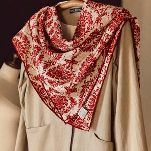 Peut inclure: Un foulard carré arborant un motif damassé rouge vif sur fond crème, drapé sur un vêtement beige à manches longues. Le foulard présente une bordure rouge foncé et une petite étiquette noire près du col portant l'inscription VOGUE.