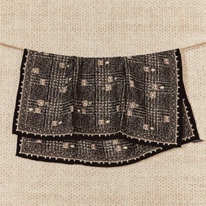 May include: A black and white houndstooth patterned scarf featuring a decorative border with intricate white embroidery and small, scattered metallic embellishments throughout the fabric, hanging horizontally from a thin string.