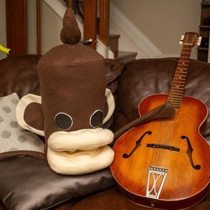 May include: A brown and cream-colored plush monkey toy with a cylindrical head, large ears, and circular eyes, positioned next to a hollow-body acoustic guitar with a sunburst finish, f-holes, and a metal tailpiece on a dark brown leather surface.