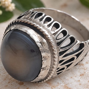 May include: A silver ring featuring a large, smooth, round, grey agate stone set in a bezel. The band is decorated with a twisted rope pattern and an openwork design consisting of elongated, teardrop-shaped cutouts around the circumference.