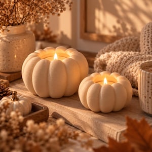 Peut inclure: Deux bougies blanc cassé en forme de citrouille avec des textures côtelées et des mèches allumées. Les bougies sont disposées sur une surface en bois avec une petite citrouille blanche, un vase en céramique, du tissu tricoté et une tasse.