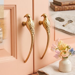 May include: Two brass cabinet handles shaped like birds with long, curved tails. The metallic bird figures feature detailed wing textures and are mounted vertically on a light peach-coloured cabinet door surface, providing a functional grip.