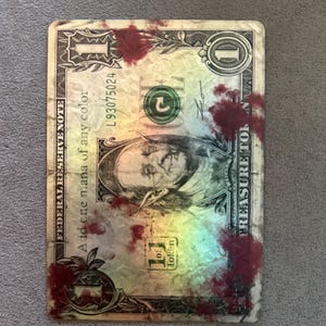May include: A rectangular game card designed to resemble a one-dollar bill with a holographic finish. The card features a central illustration of a goblin-like figure, the text 'Add one mana of any colour', '1 of 1 Token', and red splatters.