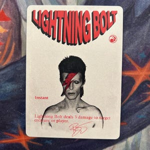 Pode incluir: Um cartão colecionável retangular com o texto LIGHTNING BOLT em vermelho no topo. Abaixo, uma ilustração em preto e branco de uma pessoa com um raio vermelho sobre o olho. O texto Instant e Lightning Bolt deals 3 damage to target creature or player aparece em vermelho, junto com uma assinatura vermelha na parte inferior.
