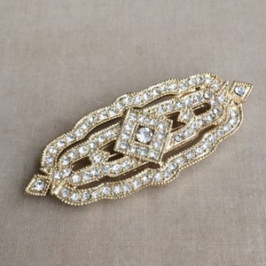 May include: A gold-toned brooch with a geometric design, featuring a central diamond-shaped cluster of clear rhinestones surrounded by smaller rhinestones.