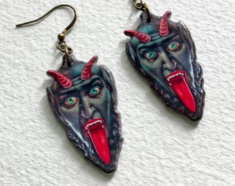 May include: A pair of dangle earrings featuring a cartoon demon face with a red tongue and horns. The earrings are made of metal and have a bronze finish.