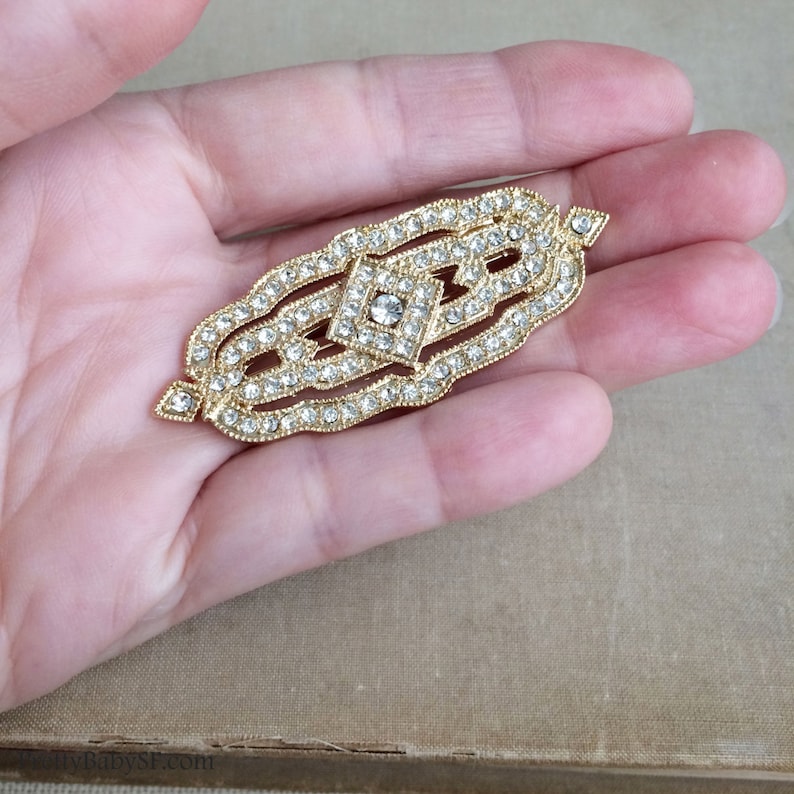 May include: A gold-toned brooch with a geometric design, featuring a central diamond-shaped rhinestone surrounded by smaller rhinestones.