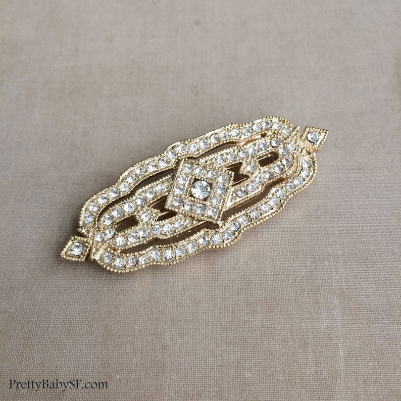 May include: A gold-toned brooch with a geometric design, featuring a central diamond-shaped cluster of clear rhinestones surrounded by smaller rhinestones.
