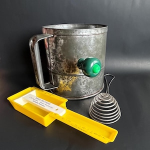 May include: A collection of vintage kitchen tools, including a metal flour sifter with a green handle, a yellow measuring tool with text, and a wire whisk. The sifter has a handle and a green knob. The measuring tool has a white ruler.