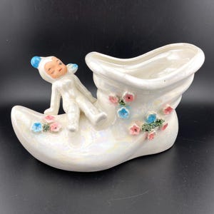 May include: A white ceramic planter shaped like a shoe with a fairy figurine sitting on top. The planter is decorated with pink and blue flowers.