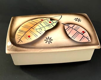May include: A rectangular ceramic trinket box with a lid. The lid is decorated with two stylized feather designs in shades of orange and pink, along with small black flower motifs. The box has a tan base and a brown rim.