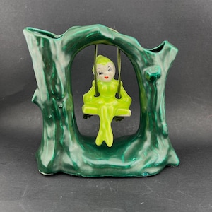 May include: A vintage ceramic vase in the shape of a tree trunk, painted in shades of green. A small figurine of a seated elf in lime green attire is suspended from the tree's archway. The elf has rosy cheeks and red lips.