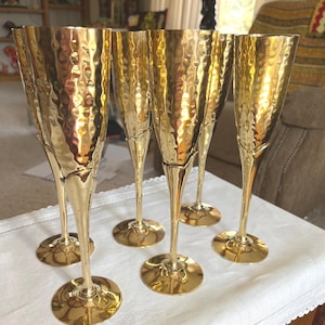 May include: Set of six gold-colored champagne flutes. Each flute has a textured, hammered finish on the bowl and a slender stem leading to a round base. The flutes are arranged on a white tablecloth, reflecting light.
