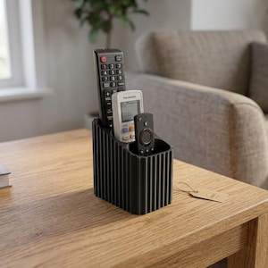 May include: A black, rectangular remote control holder with a vertical ribbed texture. The organizer features three distinct compartments containing a tall black remote, a white Panasonic remote, and a small black streaming device remote.