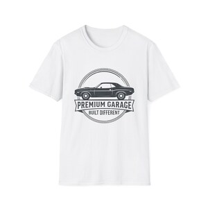 May include: A white short-sleeve t-shirt featuring a black graphic print on the chest. The design shows a side profile of a classic muscle car enclosed in a circular rope border, with the text PREMIUM GARAGE and BUILT DIFFERENT below it.