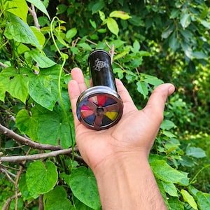 May include: A cylindrical black kaleidoscope featuring a circular viewing end with a metal frame divided into six segments, each displaying a different color including red, yellow, blue, and pink, with a small white graphic printed on the side.