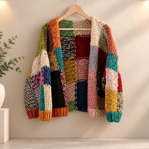 Puede incluir: Un cárdigan de punto grueso con un diseño de parches que presenta grandes cuadrados en varios colores, incluyendo naranja, verde, negro, amarillo, rosa, rojo y azul cerceta. La prenda cuelga de una percha de madera y tiene el texto StoreByElvara.