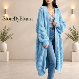Puede incluir: Un cárdigan de punto azul claro de manga larga, con frente abierto, textura acanalada y dos bolsillos delanteros. La prenda se lleva sobre una camiseta de tirantes gris y vaqueros azules. El texto "StoreByElvara" aparece arriba a la izquierda.