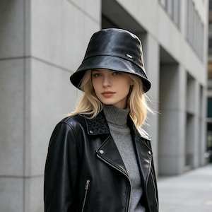 Puede incluir: Un sombrero de pescador negro con acabado liso y un pequeño detalle de logotipo metálico en el lateral. El sombrero presenta un ala ancha inclinada hacia abajo y una copa estructurada, lo que proporciona un aspecto limpio y uniforme a este accesorio.