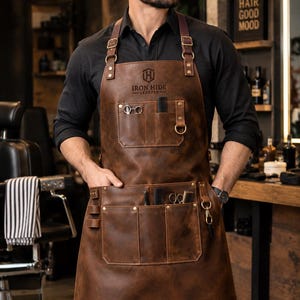 Puede incluir: Un delantal de cuero marrón con el logotipo de Iron Hide Leather, correas ajustables con hebillas metálicas y múltiples bolsillos frontales que contienen herramientas de peluquería como tijeras y peines. El cuero tiene un acabado oscuro y desgastado.