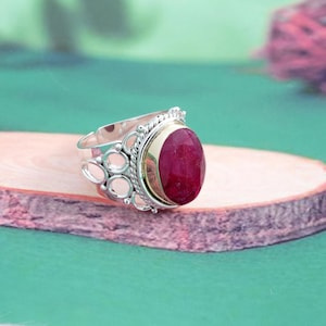 May include: A silver ring featuring a large, oval-shaped, deep red gemstone set in a bezel. The band is crafted with intricate openwork circular patterns and a twisted rope-style border surrounding the central stone, creating a detailed design.