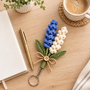 Peut inclure: Un porte-clés en crochet représentant deux tiges de fleurs en bleu et crème, accompagnées de deux feuilles vertes. Les tiges sont entourées de fil vert, nouées avec un nœud en ficelle brune et ornées d'un petit bouton en bois.