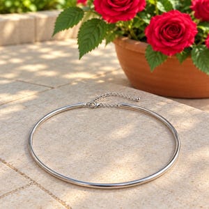 Peut inclure: Un collier ras-du-cou en métal argenté composé d'un anneau rigide et lisse. Le collier est doté d'une courte chaînette d'extension réglable terminée par une petite perle sphérique, le tout fixé par un fermoir mousqueton.