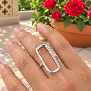 Può includere: Un anello color argento caratterizzato da un grande design a cornice rettangolare aperta. L'anello è indossato su un dito, mettendo in risalto la superficie metallica liscia e lucida della forma geometrica che si estende verticalmente sul dito.